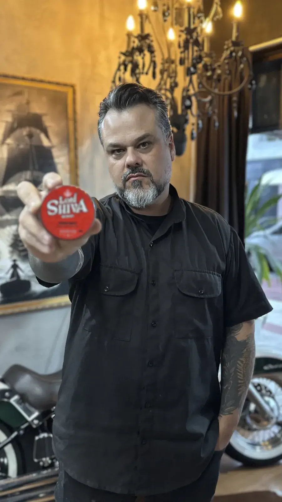 Homem com barba estilizada em ambiente de barbearia clássica segurando a pomada Power Matte em destaque para a câmera.