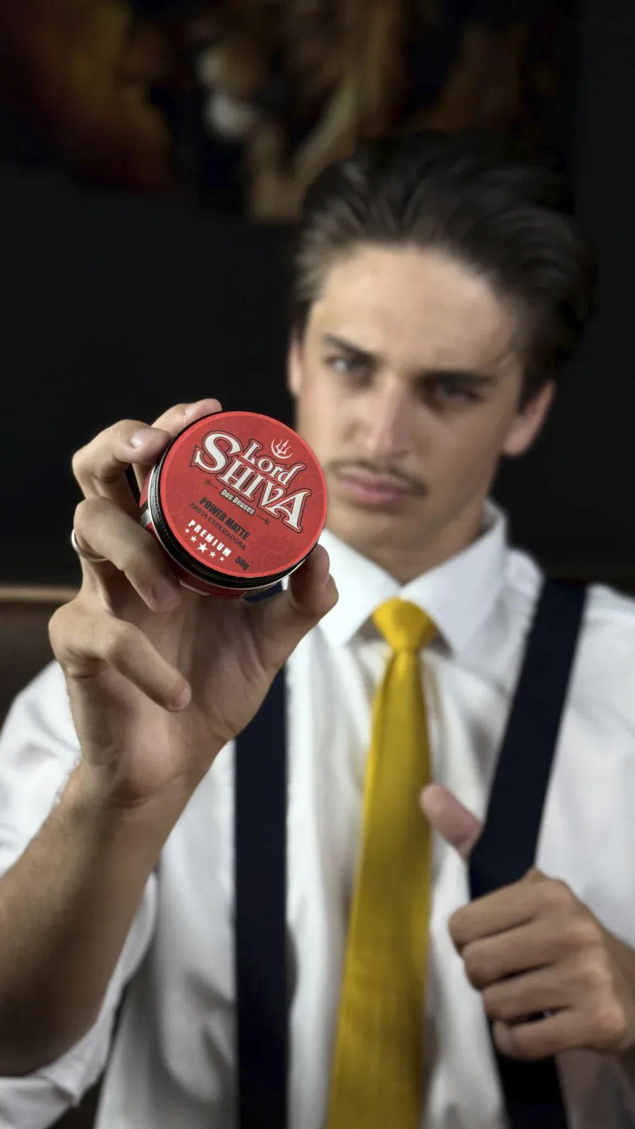 Close-up das mãos de um modelo segurando o pote vermelho da pasta estilizadora Power Matte da Lord Shiva, com foco no produto.