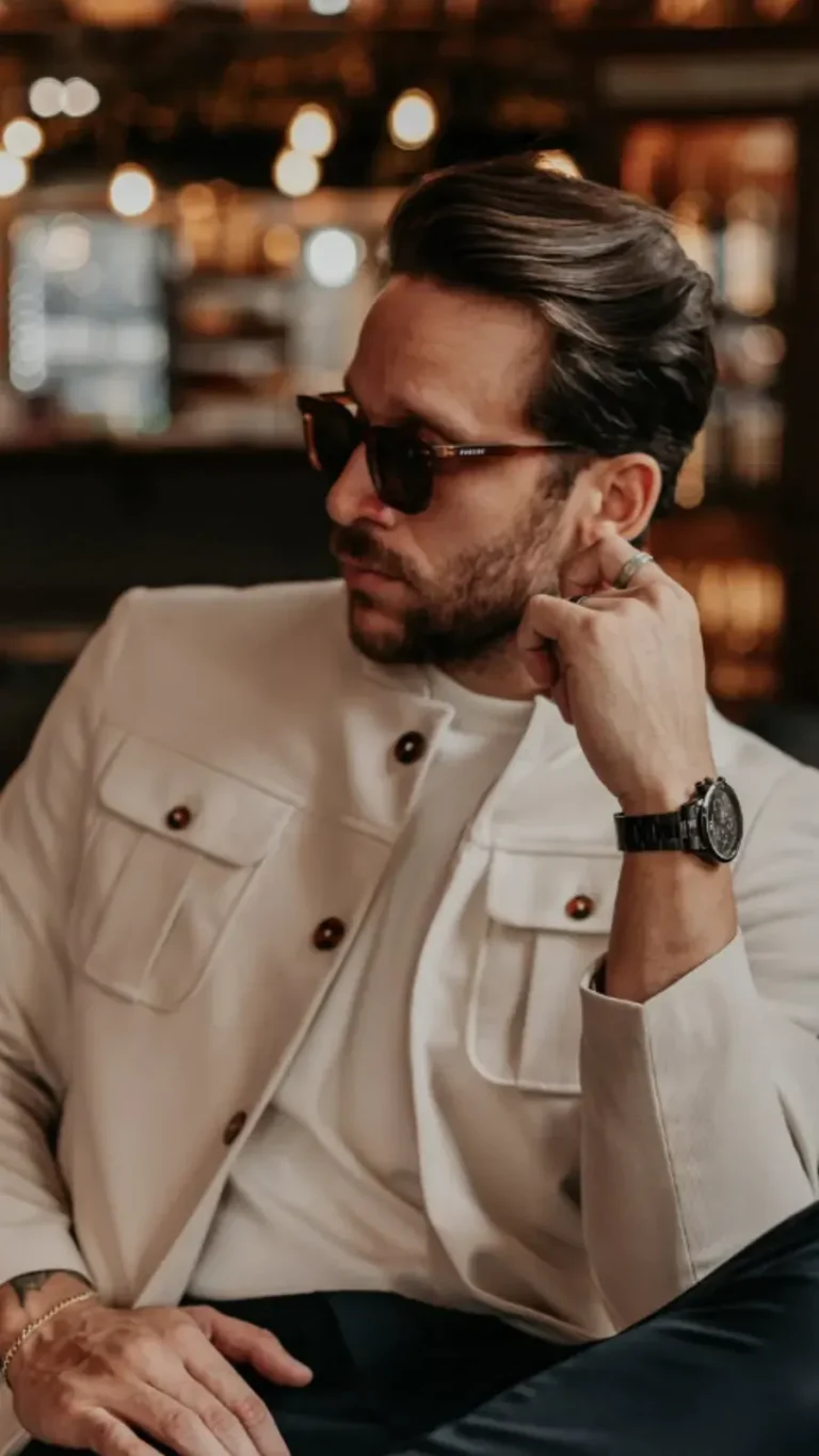 Homem elegante com jaqueta bege e óculos escuros, sentado em um ambiente interno refinado.