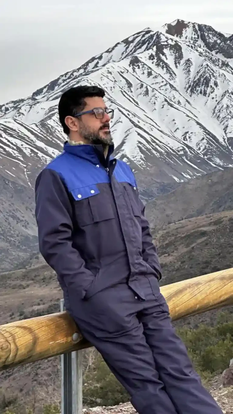 Homem de óculos de grau e jaqueta azul marinho olhando para o horizonte com montanhas nevadas ao fundo.