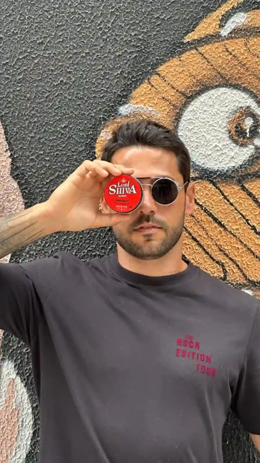 Homem segurando o pote da pomada Power Matte sobre um dos olhos, em frente a uma parede grafitada.