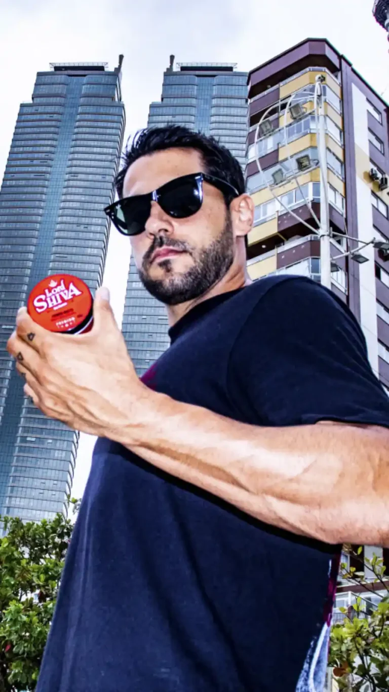 Homem de óculos escuros em ângulo baixo segurando a pomada Power Matte contra um fundo de arranha-céus modernos.