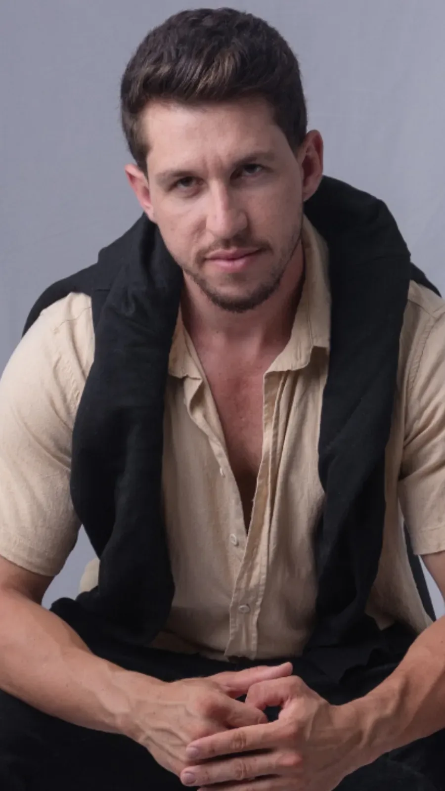 Homem sentado com camisa bege e blusa preta sobre os ombros, olhando para a câmera com cabelo modelado.